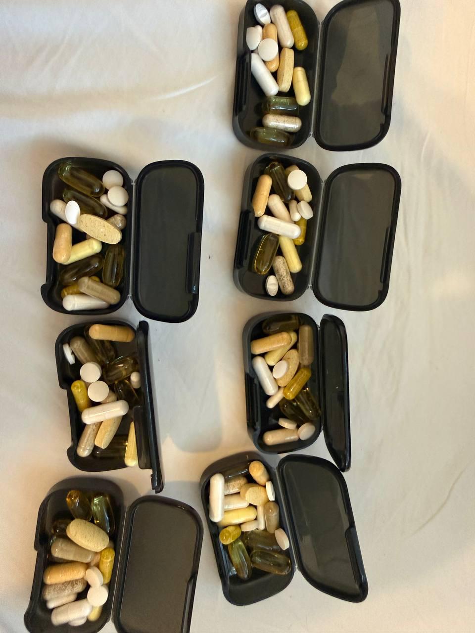 Seven open black pill boxes on a white sheet, each holding a mix of capsules and tablets in different colours.