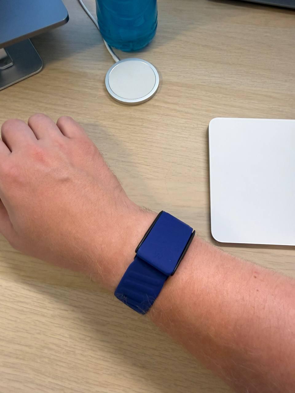 A WHOOP band with a blue strap on a wrist, a wireless charger and a trackpad on the desk; the band has no screen.